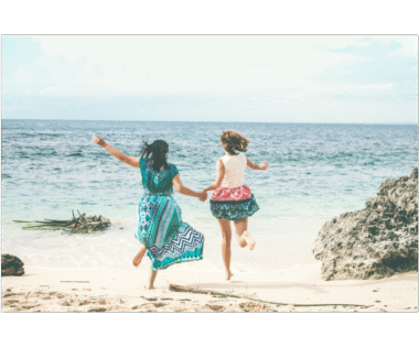 Two women holding hands and joyfully jumping on a sandy beach by the ocean, symbolizing vitality, connection, and freedom in midlife.