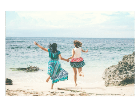 Two women holding hands and joyfully jumping on a sandy beach by the ocean, symbolizing vitality, connection, and freedom in midlife.