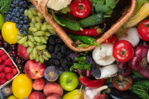 Colourful assortment of fresh fruits and vegetables, including tomatoes, grapes, peppers, berries, and greens spilling from a wicker basket.