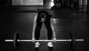 Woman in a gym preparing to lift a barbell, wearing athletic clothing and sneakers, in a black-and-white photo emphasizing strength and focus.
